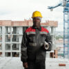 Mature African American builder with rolled blueprint looking at camera while standing against unfinished buildings at construction site